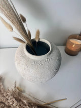 A textured light gray concrete-style ceramic vase with dried pampas grass stems inside, styled on a white surface with a blurred amber candle in the background.