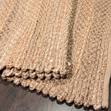A close-up of a textured woven area rug in a warm taupe-beige color, showing its chunky braided pattern and a knotted fringe edge, laid over a dark wood floor. Callista Home