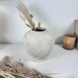 A textured, round off-white ceramic vase filled with dried pampas grass, styled on a white surface next to a lit amber glass candle.