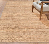 A natural jute area rug in a warm tan color, shown in a lifestyle setting on a light wood floor next to a wooden armchair with a light beige cushion. Callista Home