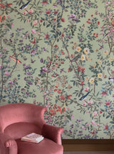A pink upholstered armchair in a styled interior setting, with a book resting on the seat and a wall covered in dense floral and bird wallpaper behind it. Callista Home