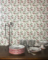 A tabletop display featuring a stack of white ceramic plates with red and green floral patterns, several clear drinking glasses, silver-toned cutlery, and a white pedestal bowl, set against a wall covered in matching floral wallpaper. Buisson de Roses 58A French floral wallpaper by Antoinette Poisson, inspired by historic domino paper and printed in Brittany, France. Callista Home