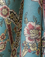 A close-up of a patterned fabric with a turquoise blue background, featuring large white and pink flowers, yellow accents, and dark outlines. Callista Home