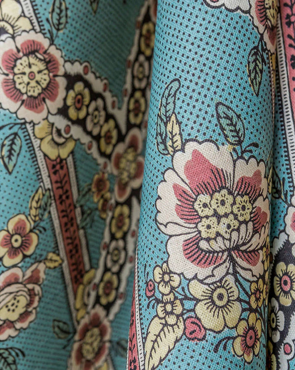 A close-up of a patterned fabric with a turquoise blue background, featuring large white and pink flowers, yellow accents, and dark outlines. Callista Home