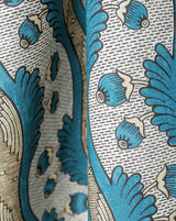 Close-up of a patterned fabric with blue stylized floral or leaf motifs, beige outlines, and a white background with fine gray herringbone lines. Callista Home, Grenades 2A botanical pomegranate French linen fabric