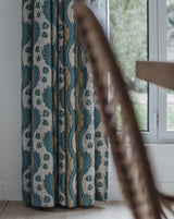 A close-up of patterned curtains hanging in front of a window, with a blurred wooden chair in the foreground and a bright outdoor view visible on the right. Grenades 2A botanical pomegranate French linen fabric Callista Home