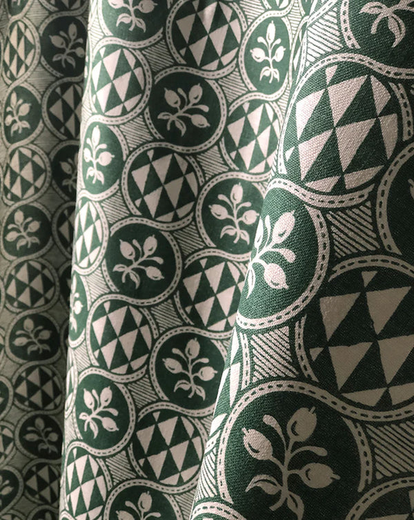 A close-up of patterned fabric with a repeating circular medallion design in dark green, cream, and gray, shown with soft folds. Olives 55A botanical French linen fabric by Antoinette Poisson Callista Home