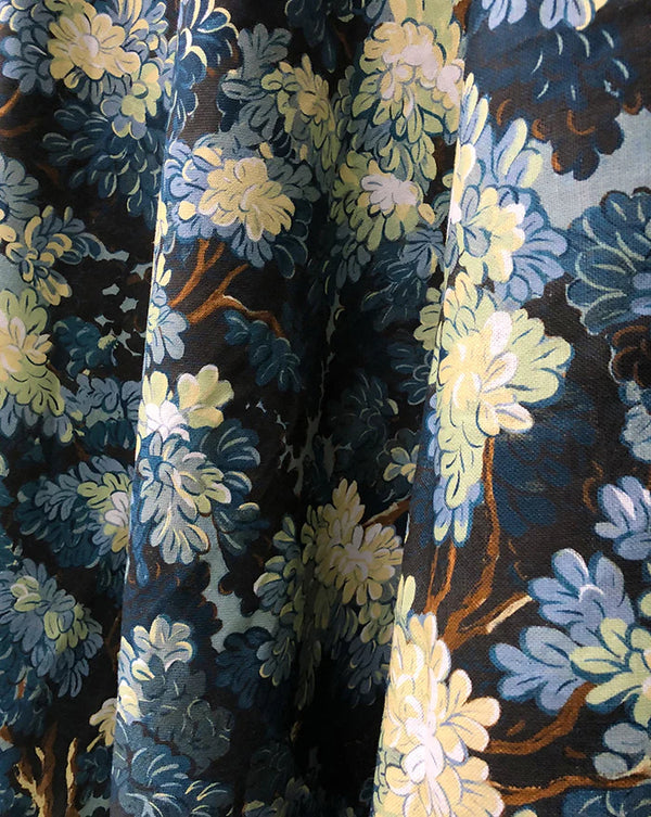 Close-up of a draped fabric with a dense floral pattern in navy blue, light blue, pale yellow, and brown on a dark background. Joli Bois botanical French linen fabric by Antoinette Poisson Callista Home